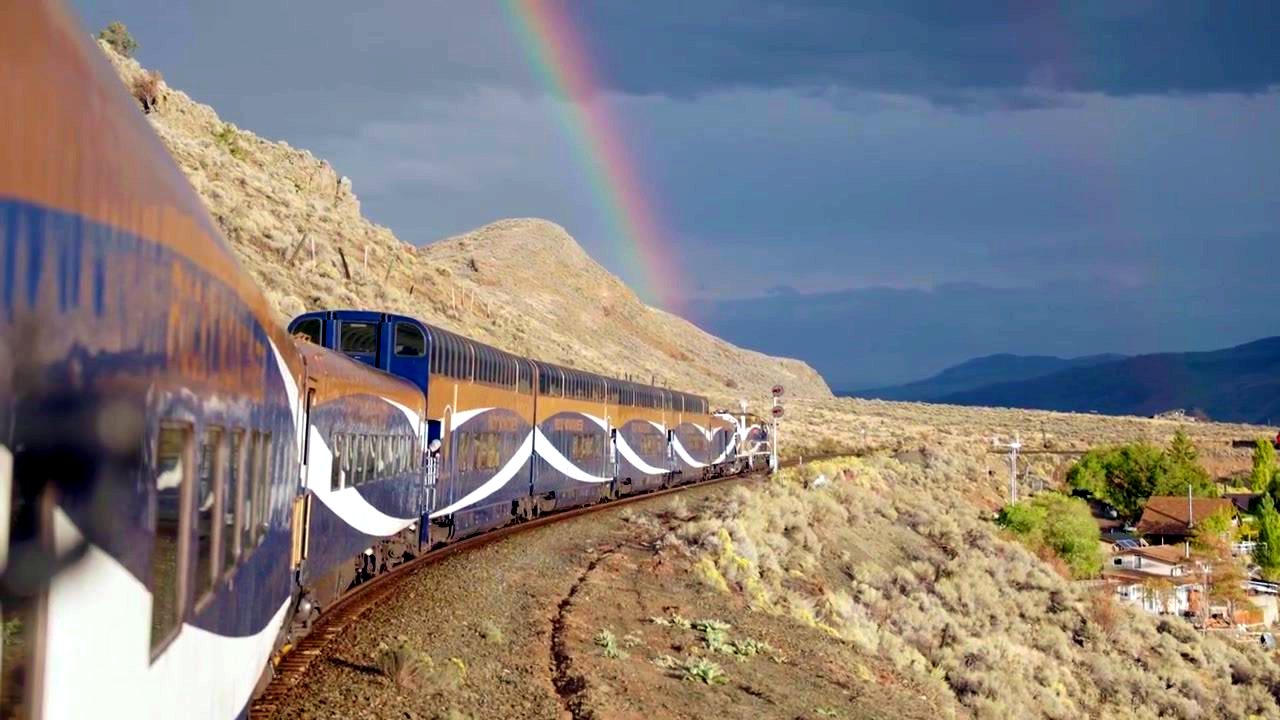 The Rocky Mountaineer winding along a sunlit hillside beneath a rainbow