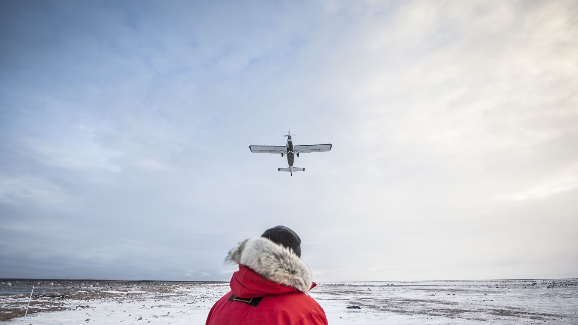 A visitor in a fur-trimmed parka watching a small plane fly over the snowy shore