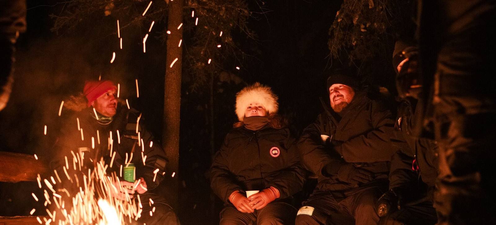 Two guests in winter gear beside a crackling fire at night