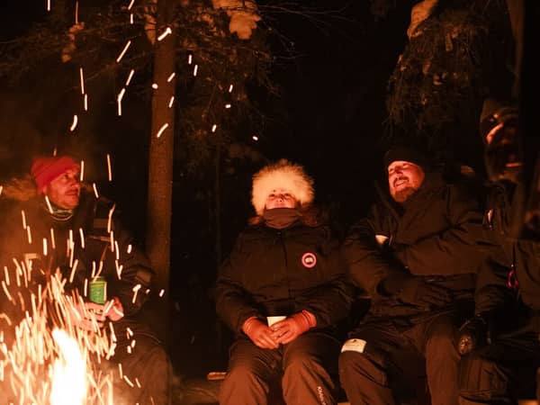 Two guests in winter gear beside a crackling fire at night