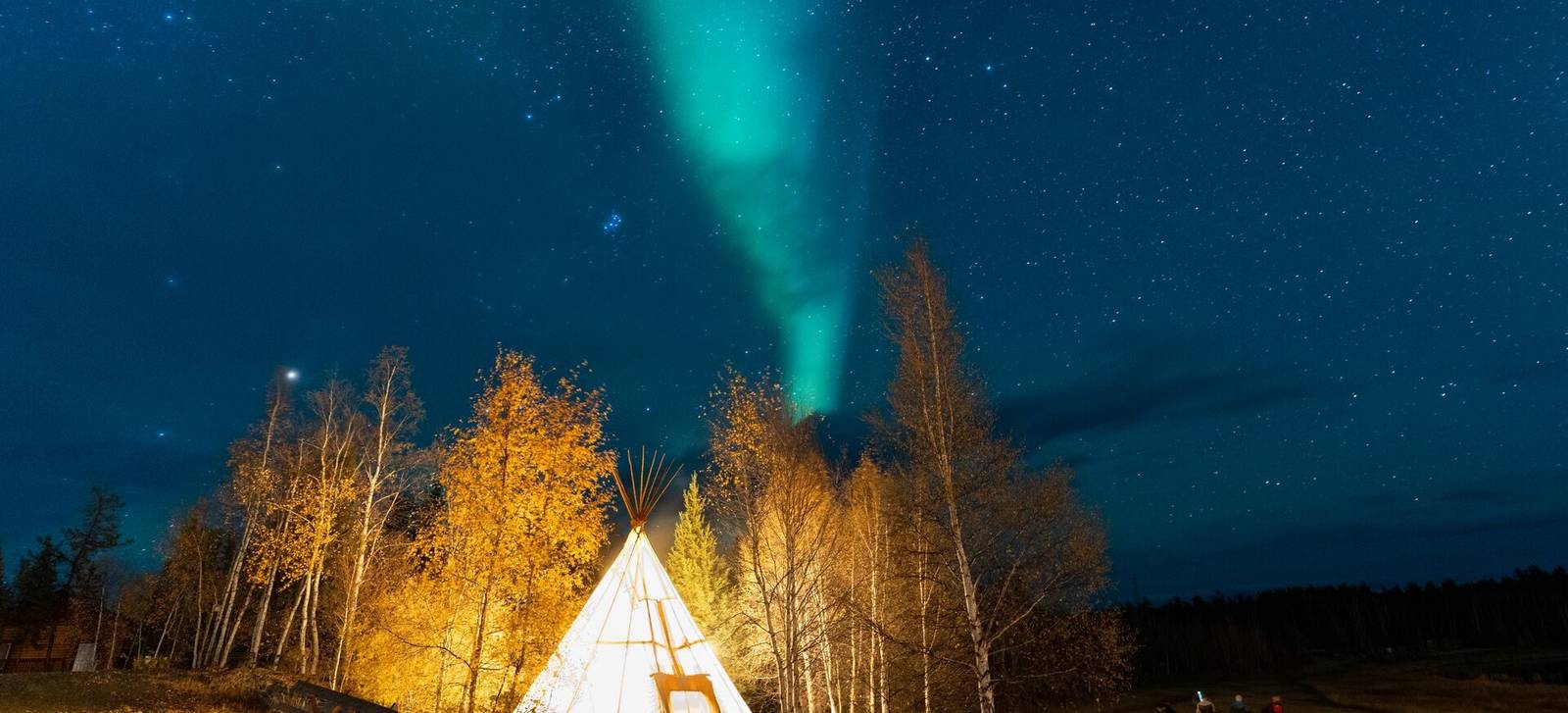A glowing tipi among golden trees under a night sky streaked with aurora