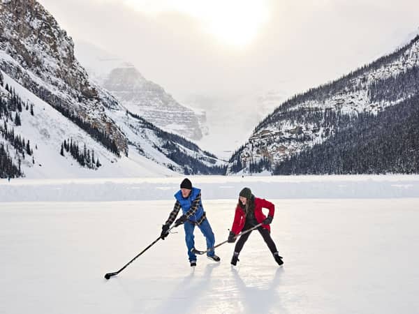 Two skaters with hockey sticks on frozen Lake Louise beneath snowy peaks